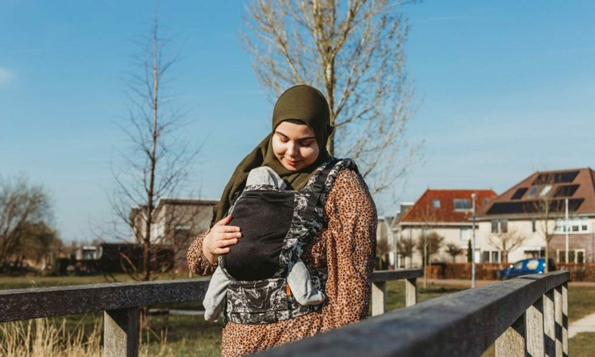 Feride met haar baby in een Tula draagzak bij een houten hek, buiten in de zon