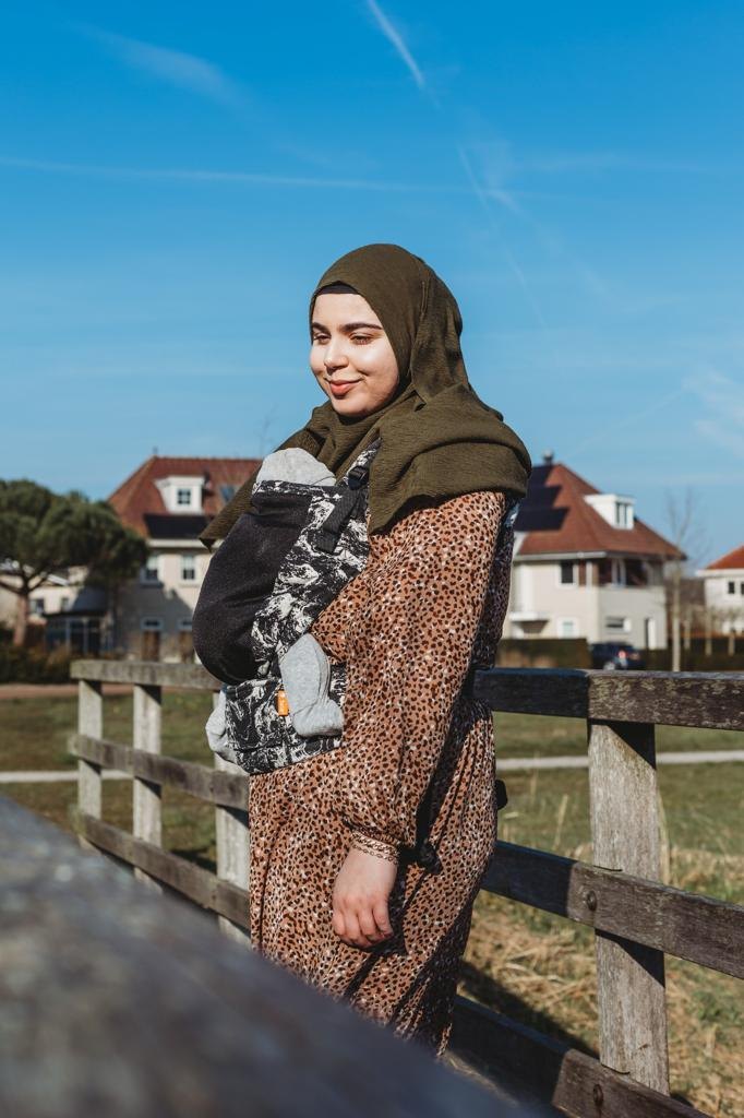 Feride staat glimlachend bij een houten hek met haar baby in een Tula draagzak