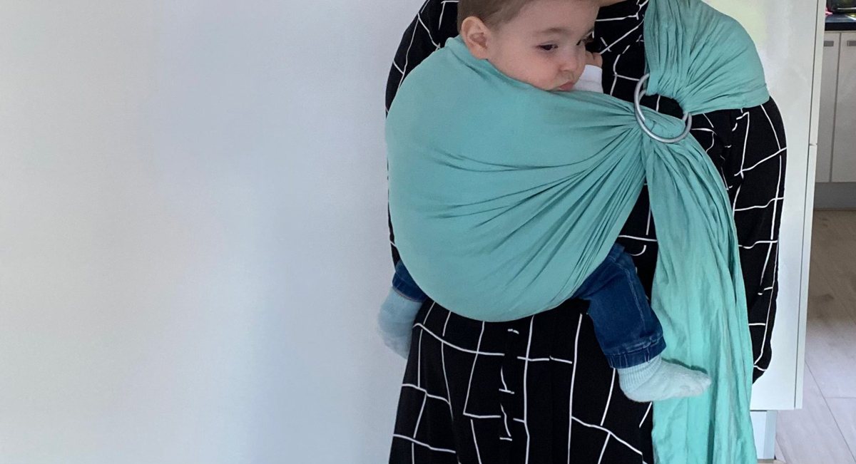 Kindje in een turquoise ringsling op de heup, binnenshuis tegen een lichte muur